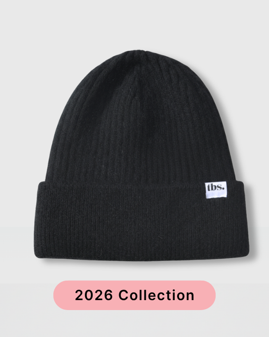 A black ribbed knit beanie hat with a small white 'tbs.' logo patch on the cuff, shown against a light gray background with a pink '2026 Collection' label below. silk lined beanie hat - prevents frizz and static as well as breakage.