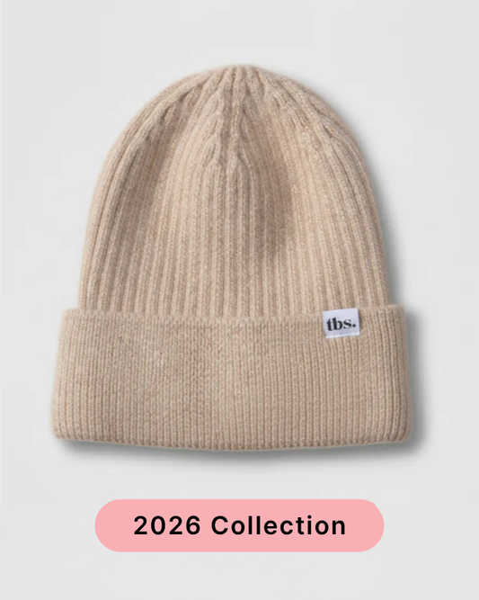 A beige ribbed knit beanie hat with a folded cuff and a small white 'tbs.' label on the side, shown on a light gray background with a pink '2026 Collection' banner at the bottom. silk lined beanie to prevent frizz and static.