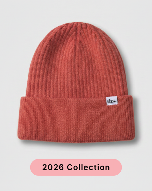A ribbed knit beanie in a muted terracotta or burnt orange color, featuring a small white 'tbs.' logo patch on the folded cuff. silk lined beanie.