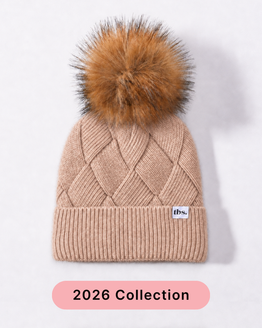A beige cable-knit beanie with a large brown faux-fur pom-pom and a small white 'tbs.' label on the cuff, shown against a light gray background with a pink '2026 Collection' banner at the bottom. silk lined.