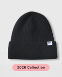 A black ribbed knit beanie hat with a small white 'tbs.' logo patch on the cuff, shown against a light gray background with a pink '2026 Collection' label below. silk lined beanie hat - prevents frizz and static as well as breakage.
