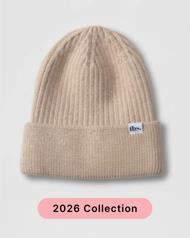 A beige ribbed knit beanie hat with a folded cuff and a small white 'tbs.' label on the side, shown on a light gray background with a pink '2026 Collection' banner at the bottom. silk lined beanie to prevent frizz and static.
