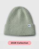A light sage green ribbed knit beanie hat with a folded cuff and a small white 'tbs.' label on the side, shown against a light gray background with a pink '2026 Collection' banner at the bottom. silk lined