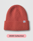 A ribbed knit beanie in a muted terracotta or burnt orange color, featuring a small white 'tbs.' logo patch on the folded cuff. silk lined beanie.